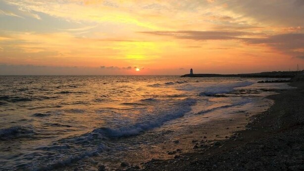 Solnedgang over en strand med bølger, en fjern fyrtårn og en stenet kystlinje.