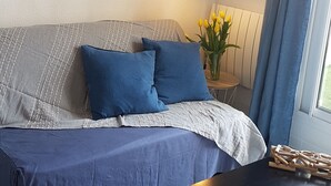 Et moderne soveværelse med en grå og blåstribet sofa, to matchende puder, et sengebord med en lampe, en vase med gule blomster og et vindue med blå gardiner.