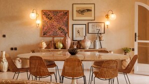 A dining area with a round table, wicker chairs, wall-mounted light fixtures, and various decorative items. - The Gardener's House (Aghouatim)