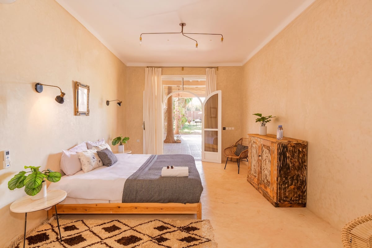 A bedroom with a bed, bedside tables, a small round table, a cabinet, and a view through an open door to an outdoor area.