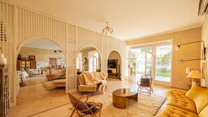 A spacious living room with a yellow sofa, a round coffee table, and a wicker chair. There are multiple archways leading to other rooms, and a large window providing natural light. - The Gardener's House (Aghouatim)