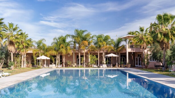 A swimming pool with lounge chairs and umbrellas, surrounded by palm trees and a building with a covered patio. - The Gardener's House (Aghouatim)