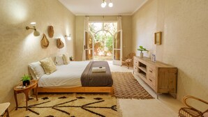 A bedroom with a bed, bedside tables, a wooden cabinet, a patterned rug, and a view of the outdoors. - The Gardener's House (Aghouatim)