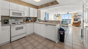 A modern kitchen with white cabinets, a microwave, stove, and dishwasher. There is a trash can and a dining area visible. - Spacious Oceanfront condo, great amenities + Free Attraction Tickets! (North Myrtle Beach)
