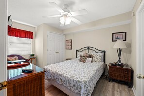 Une chambre à coucher comprenant un lit, une commode, une table de chevet, un ventilateur de plafond, une fenêtre avec des stores et une porte.