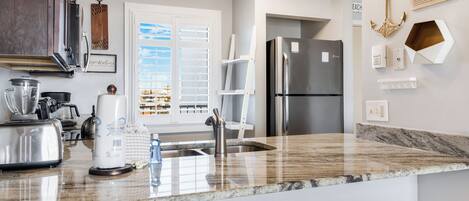 A modern kitchen with stainless steel appliances, a marble countertop, and a view through a window with blinds.