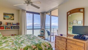 A bedroom with a bed, a dresser, a television, a bookshelf, and a view of the ocean. - Corner condo with direct beach access (Lahaina)