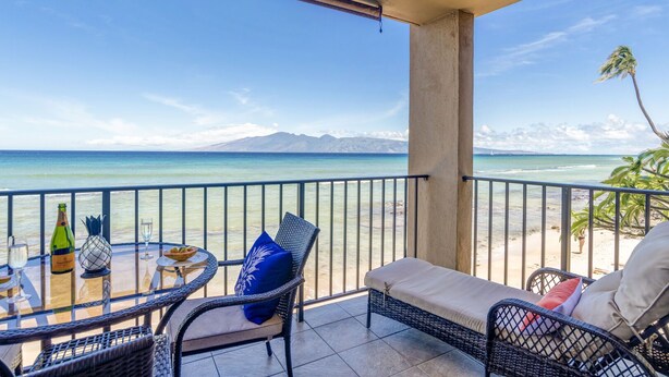 A balcony with a view of the beach and ocean, featuring wicker furniture and a table with drinks. - Corner condo with direct beach access (Lahaina)