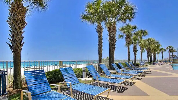 A beachside lounge area with rows of blue lounge chairs, palm trees, and a clear view of the ocean. - Gulf Front Beauty - Beach Service for 2- Owner Managed (Panama City Beach)
