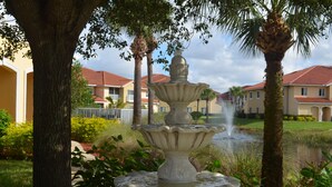 A multi-tiered stone fountain surrounded by lush greenery and palm trees, with a pond and residential buildings in the background. - Fort Myers Luxury Vacation Condo (Fort Myers)