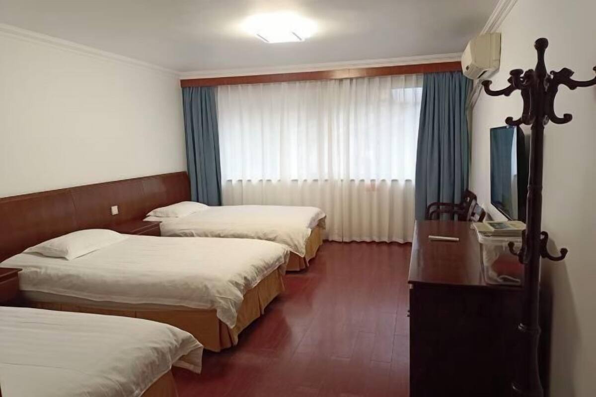 Habitación de hotel con tres camas, cabecera de madera, escritorio con espejo y ventana con cortinas.