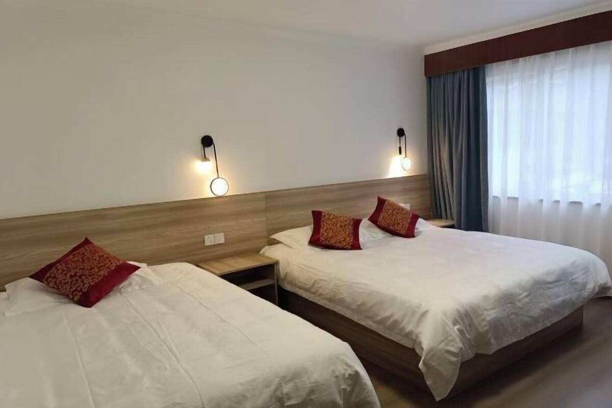 Habitación de hotel con dos camas, cabecero de madera, luces fijadas a la pared y una ventana con cortinas.