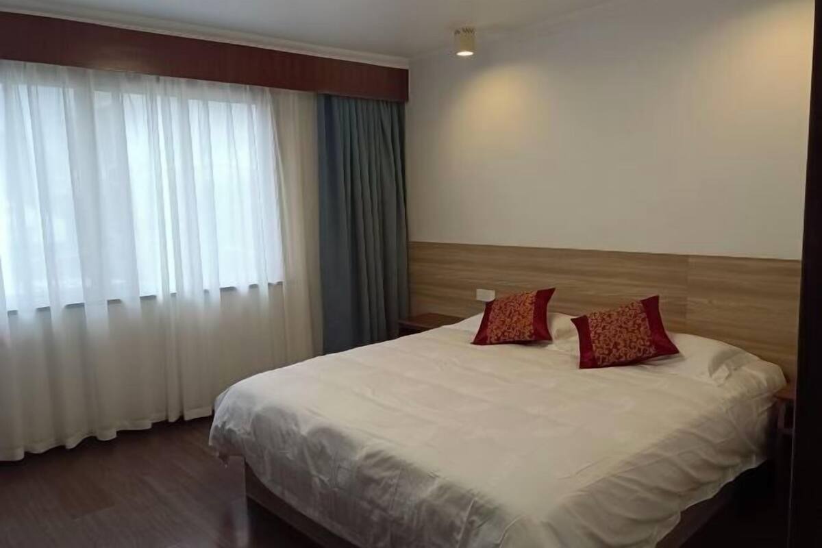 Habitación de hotel con una cama grande, dos almohadas, un cabecero de madera, una mesita de noche y una ventana con cortinas.