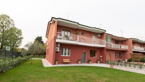 A two-story residential building with a red facade, a balcony with a white railing, and a small garden area in front. - Relax nel verde (Segrate)