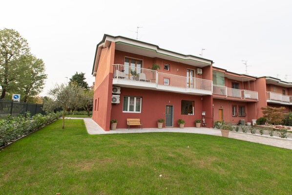 A two-story residential building with a red facade, a balcony with a white railing, and a small garden area in front. - Relax nel verde (Segrate)