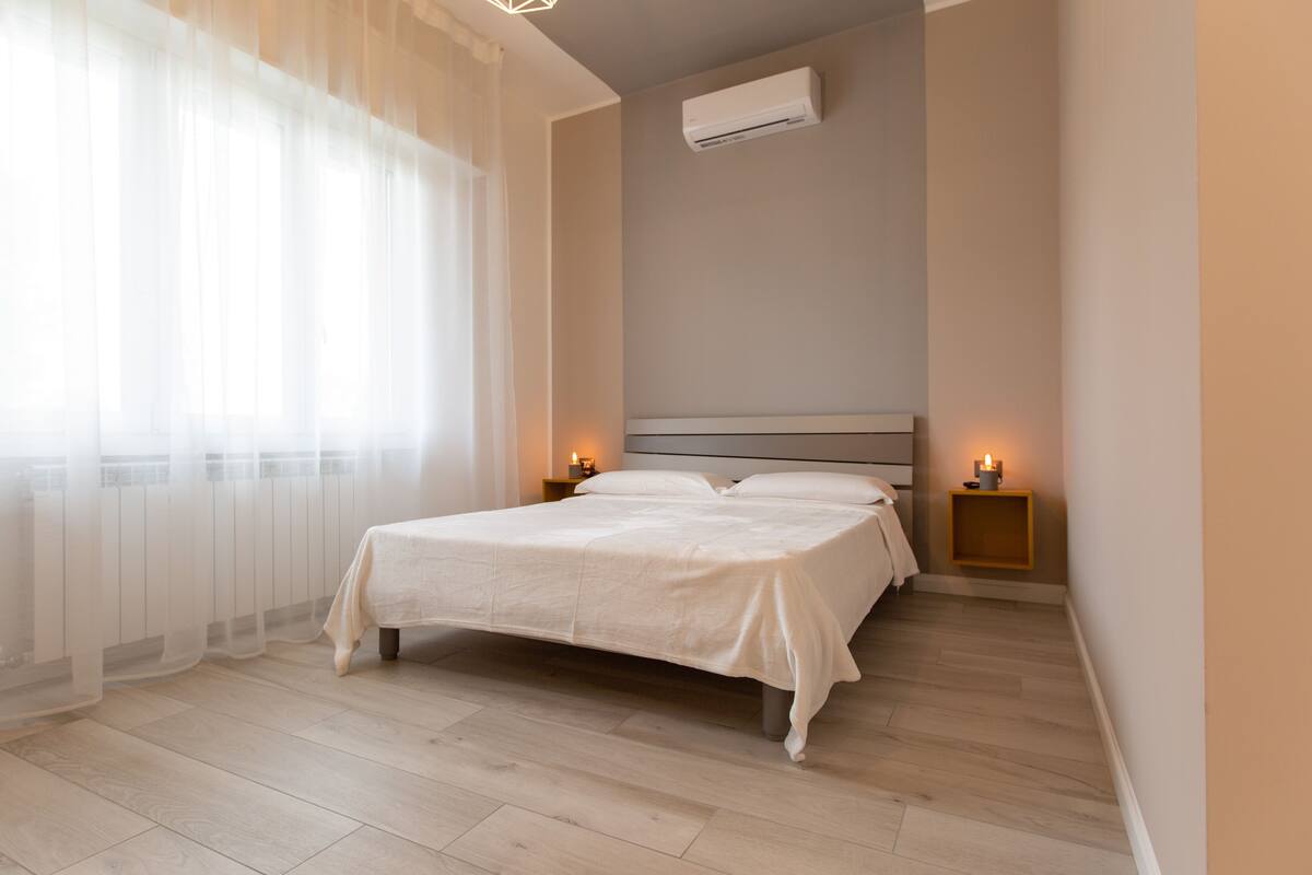Een net opgemaakt bed in een kamer met lichte houten vloeren, een witte airconditioning op de muur en twee nachtkastjes met kaarsen.