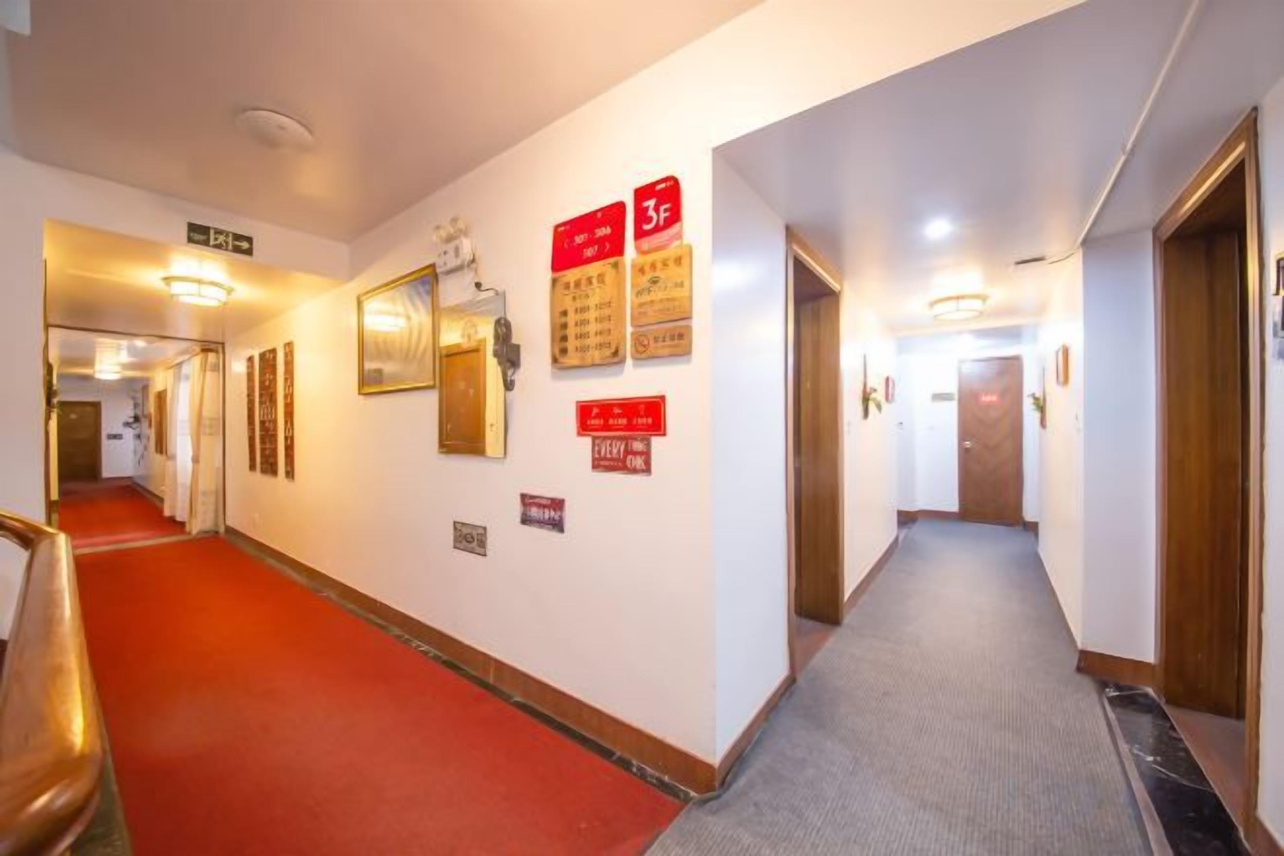 A hotel corridor with a red carpet, signs indicating room numbers, and a wooden door at the end.