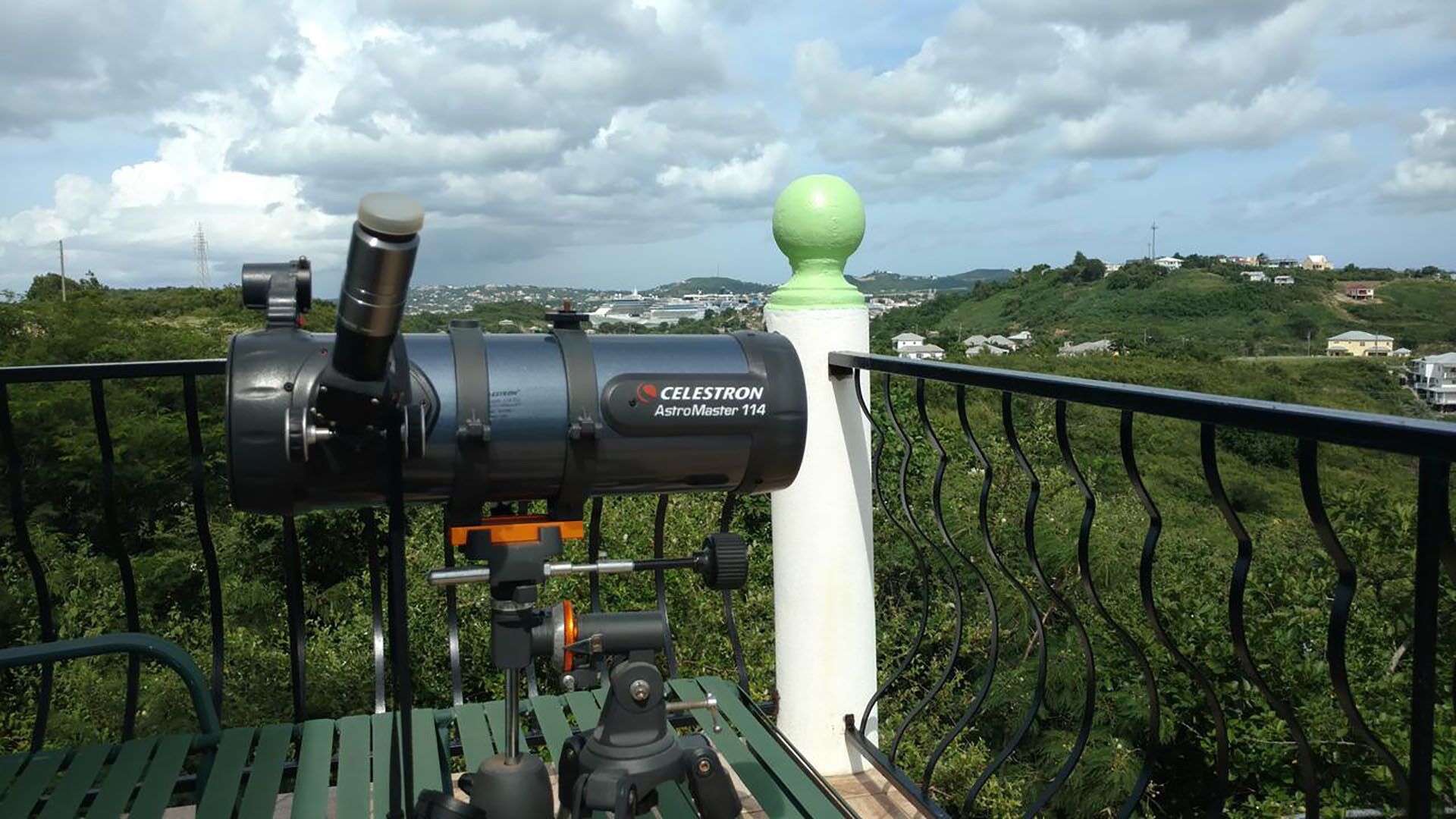 Un télescope portant la mention « CELESTRON AstroMaster 114 » est installé sur un balcon, d’où l’on peut admirer un paysage verdoyant et des bâtiments situés au loin.