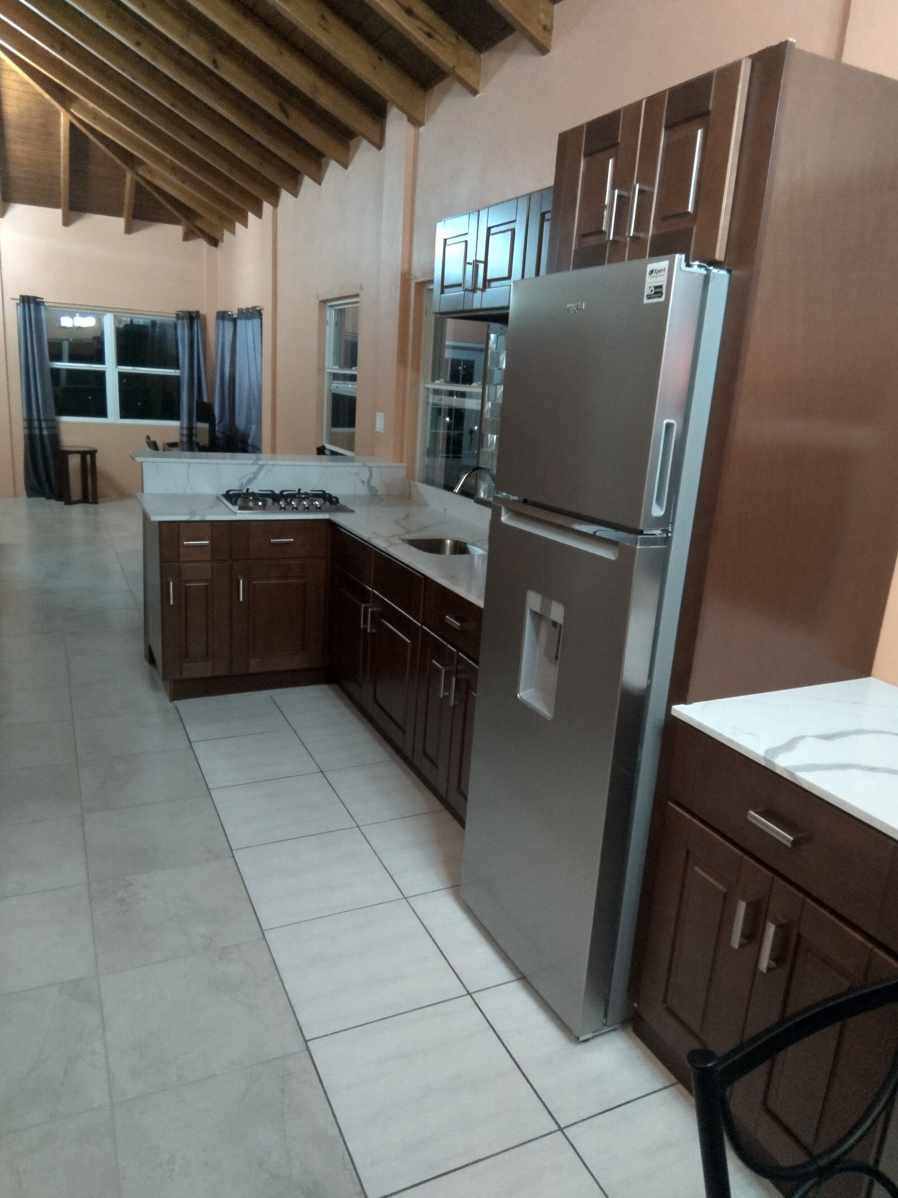 A kitchen with a stainless steel refrigerator, wooden cabinets, a marble countertop, and a tiled floor.