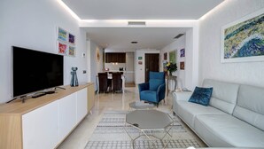 Un salon moderne avec un téléviseur à écran plat, un fauteuil bleu, un canapé blanc et un coin repas à l’arrière-plan.