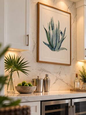 A kitchen with marble countertops, a framed botanical print, a potted plant, a bowl of limes, and a wine bottle.