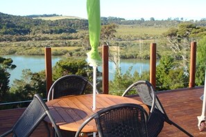 Eine Holzterrasse mit einem runden Tisch und vier Stühlen, mit Blick auf einen See und eine hügelige Landschaft.
