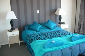 Ein ordentlich bezogenes Bett mit blauer Bettwäsche und Kissen, zwei Nachttische mit Lampen und eine gestreifte Wand im Hintergrund.