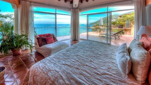 A bedroom with a large bed, a potted plant, and a view of the sea through large windows.