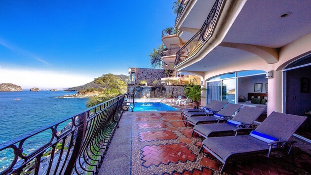 A poolside area with lounge chairs, a pool, and a view of the sea.