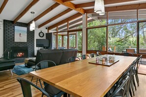 A spacious room with a large wooden table, comfortable seating, a fireplace, and large windows offering a view of the outdoors.