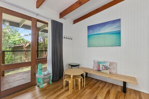 Ein Zimmer mit Holzboden, einem Esstisch mit Stühlen, einer Bank, einem Strandbild und Blick ins Freie durch eine Glastür.