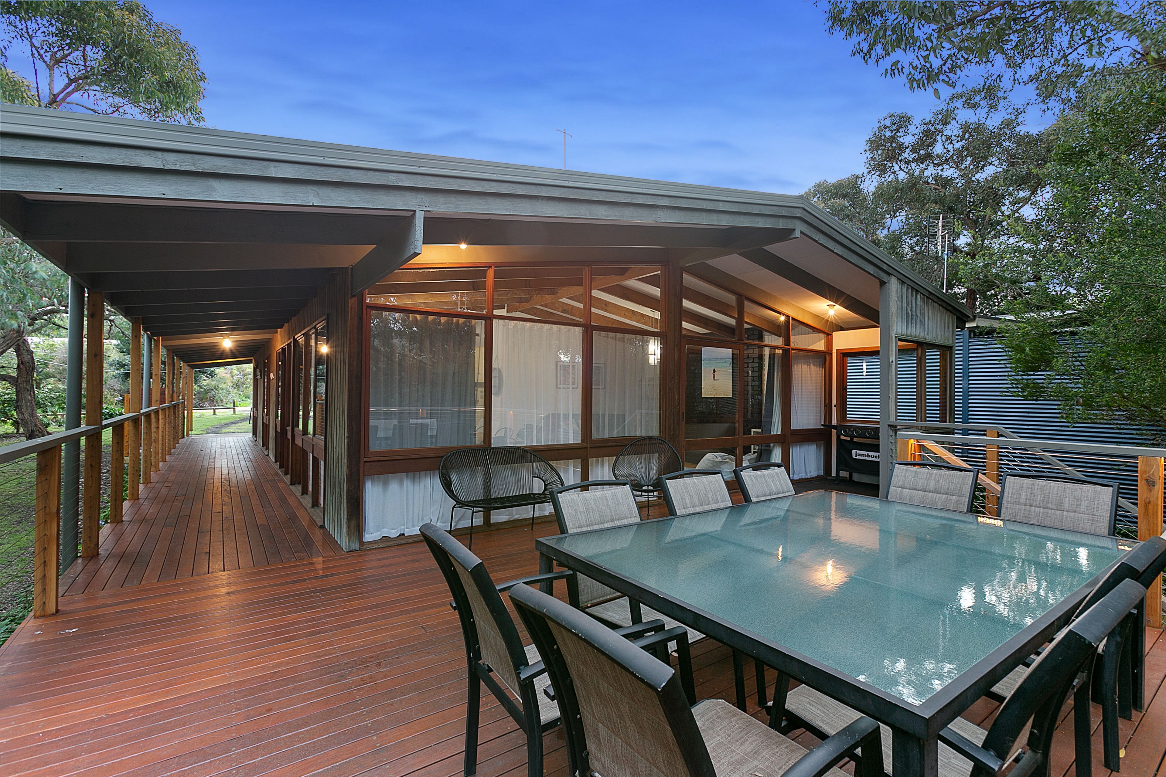 A covered outdoor deck with a glass table and chairs, surrounded by a wooden deck and a view of the beach.