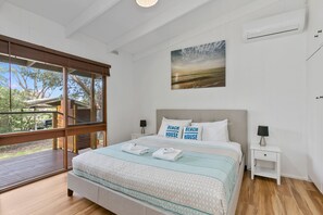 A bedroom with a large bed, two bedside tables, a window with a view of trees, and a beach scene painting on the wall.