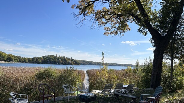 A lakeside seating area with chairs and tables, a wooden deck, and a view of a lake and forested hills. - 🌲☀️Private Lakefront prop. with Kayaks, Paddle boards, boat rental PVT dock☀️🌲 (Twin Lakes)