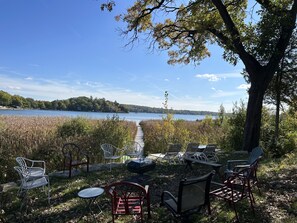 A lakeside seating area with chairs and tables, a wooden deck, and a view of a lake and forested hills. - 🌲☀️Private Lakefront prop. with Kayaks, Paddle boards, boat rental PVT dock☀️🌲 (Twin Lakes)