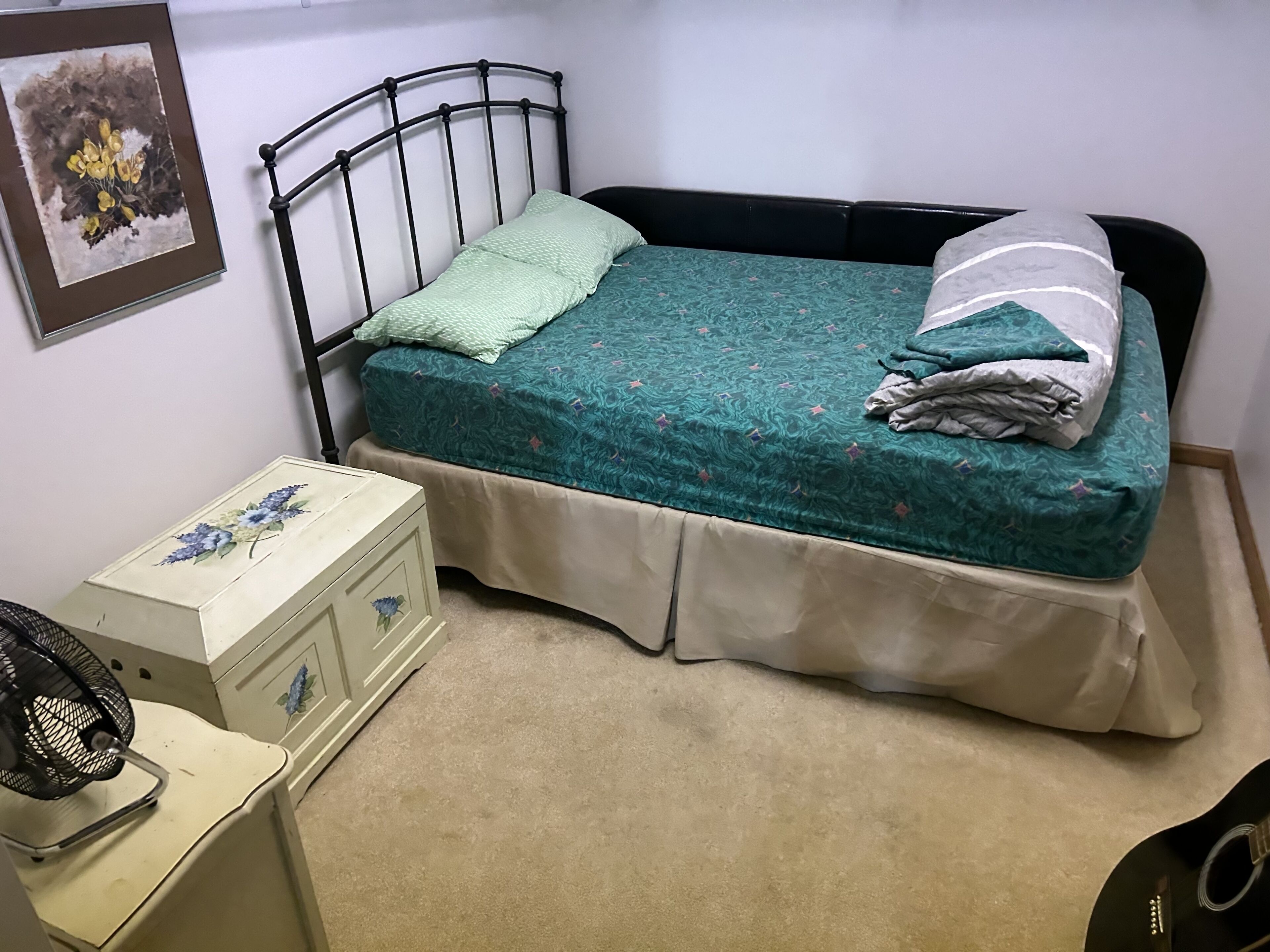 Un dormitorio pequeño y ordenado con una cama individual, una mesita de noche, un aparador y un ventilador de pared.