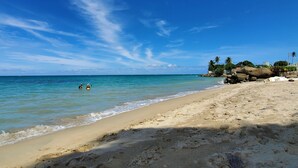 Une plage avec une eau d’un bleu limpide, un rivage sablonneux et quelques personnes dans l’eau. On aperçoit des rochers et des palmiers en arrière-plan.
