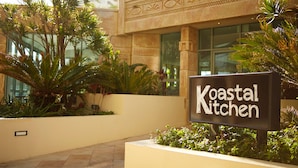 Un letrero que dice 'Koastal Kitchen' con un edificio moderno al fondo y plantas tropicales en primer plano.