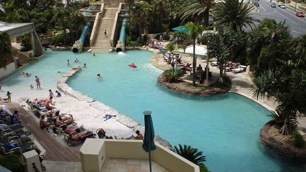 Zona de piscina en un resort con un tobogán central, varias áreas para nadar y tumbonas.