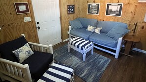 Ein gemütliches Zimmer mit Holzvertäfelung, einer weißen Tür, einem blauen Sofa, einem gestreiften Ottomanen und einem weißen Sessel mit Kissen.