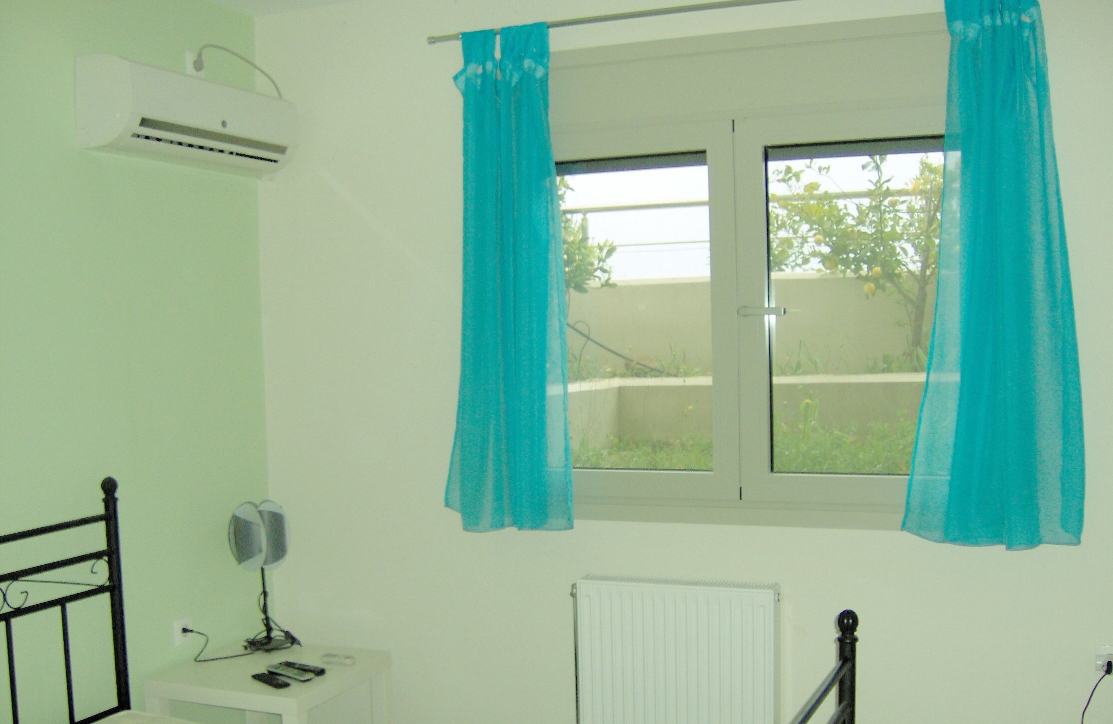A room with a bed, an air conditioner, a window with curtains, a bedside table with a lamp, and a radiator.
