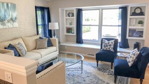 Un salon avec un mur en briques blanches, un canapé beige, un fauteuil bleu, une table basse en verre et un tapis à motifs.