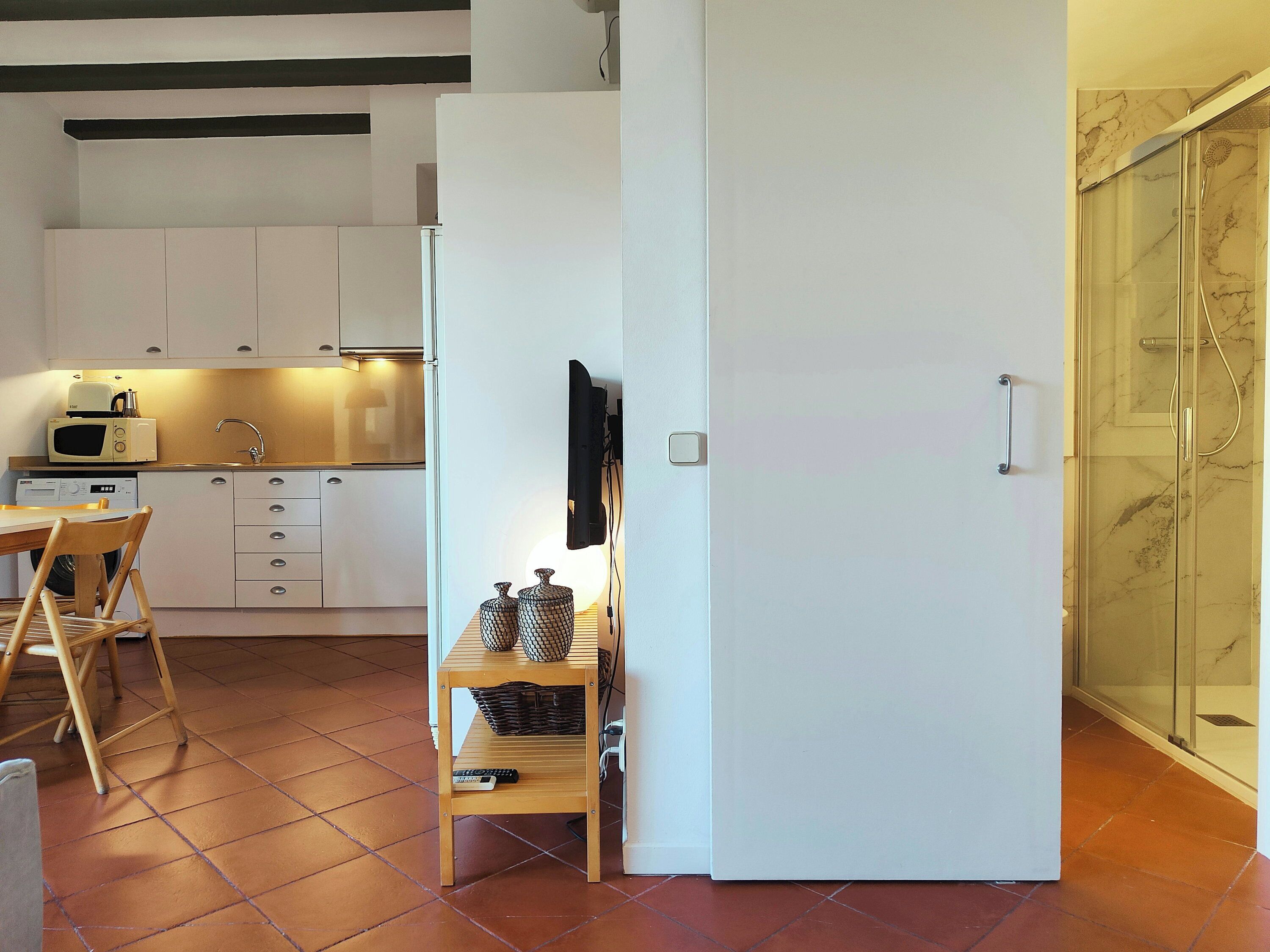 A modern kitchen with white cabinets, a microwave, and a sink. There's a wooden table with chairs and a small shelf with decorative items. A sliding door leads to a bathroom with a glass shower.