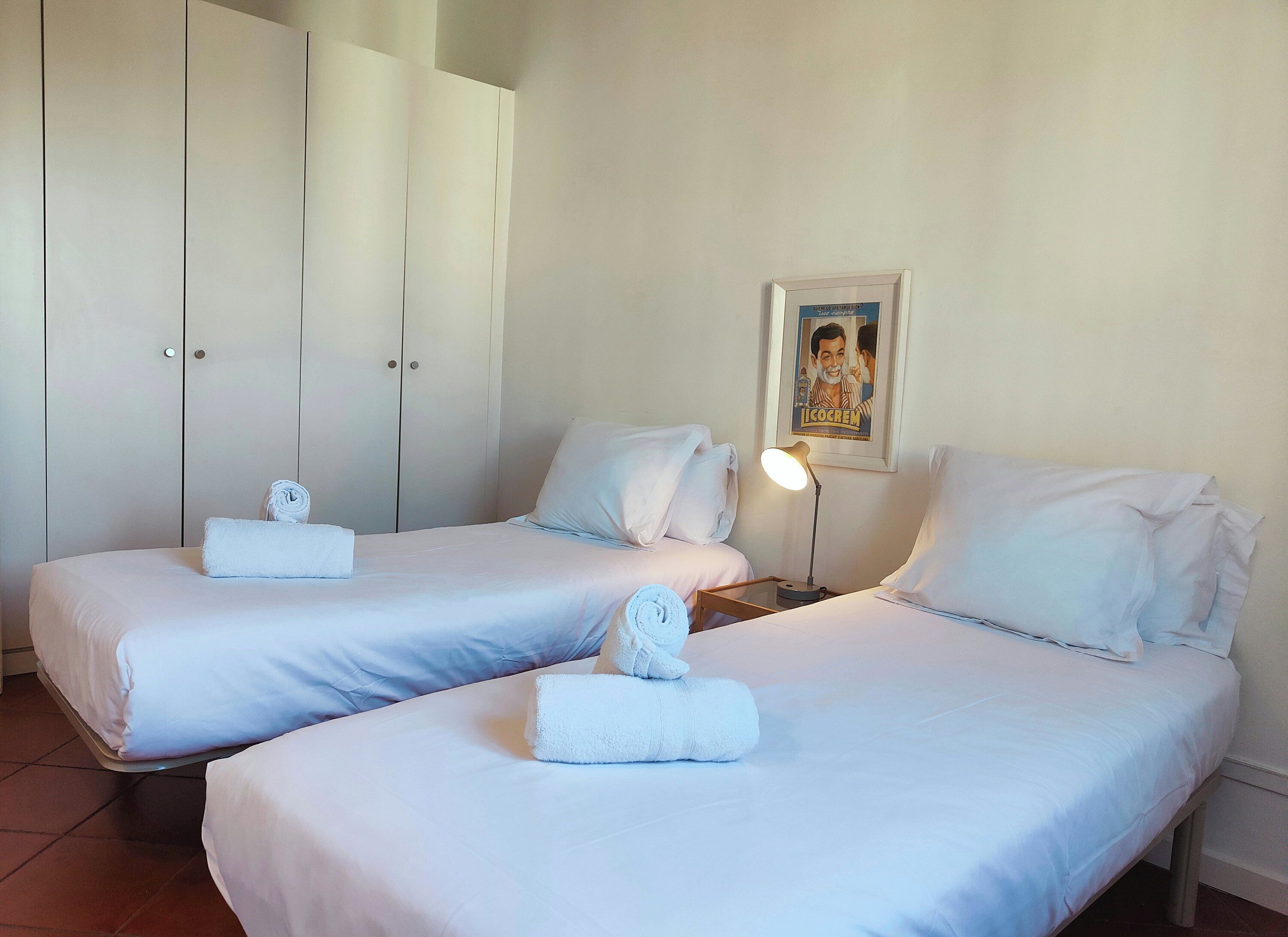 A hotel room with two beds, white linens, a nightstand with a lamp, a framed picture on the wall, and a wardrobe.