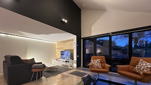 Un salon moderne avec un canapé, un fauteuil et une table basse. Un téléviseur est fixé à un mur en bois, et une porte-fenêtre coulissante donne accès à un balcon.