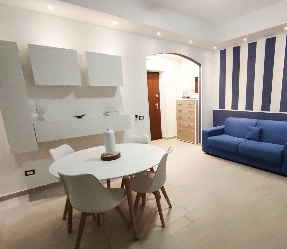 Uma sala de estar moderna com um sofá azul, uma mesa de jantar branca com cadeiras e uma parede com listras verticais.