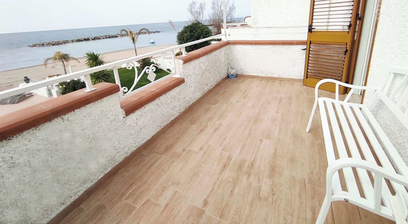 Ein Balkon mit Blick auf das Meer, weiße Stühle und eine Holztür.
