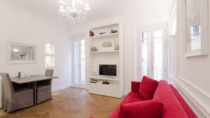 A modern living room with a herringbone floor, a red sofa, a dining table with chairs, a TV on a white entertainment unit, and a chandelier. - Luxury apartment in the heart of Rome - Spanish Steps, wi-fi, a / c, (Roma)