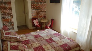 A bedroom with a brick wall, a bed with a quilt, two chairs, a small table, and a TV on the wall.