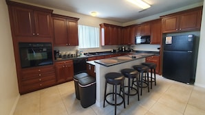 Una cocina moderna con armarios de madera, horno empotrado, microondas y refrigerador. Hay cuatro taburetes alrededor de la isla.